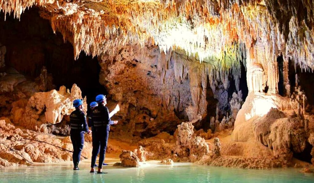 two people standing admiring an underground cenote cave with water just above their feet and looking at the top of the cave
