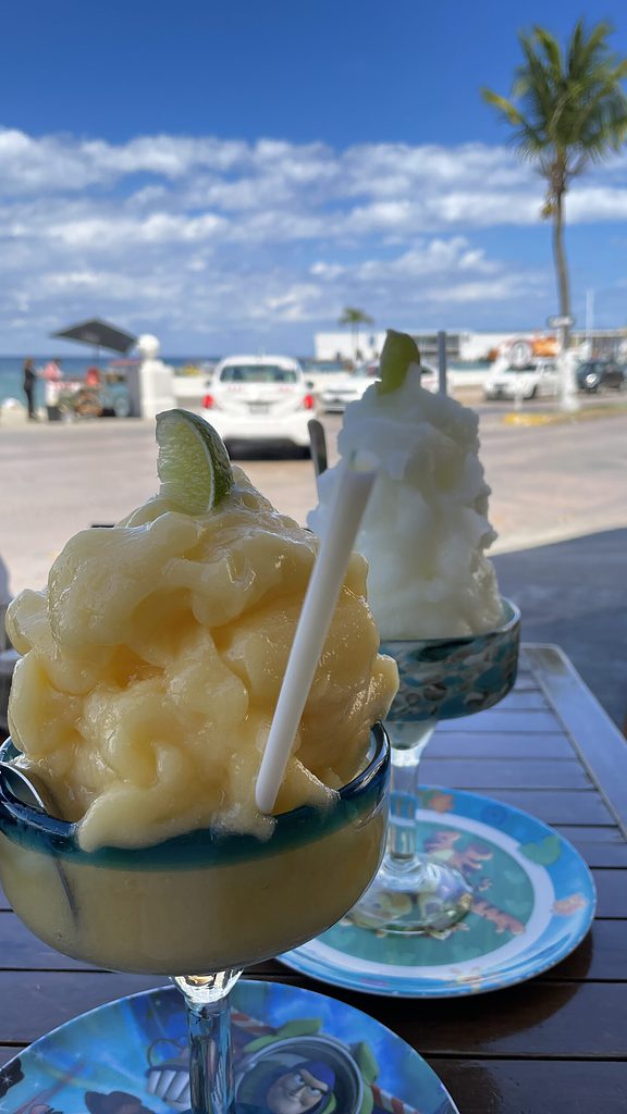 two alcoholic slushies on a table with views of ocean waters and a large cruise line ship docked in the background