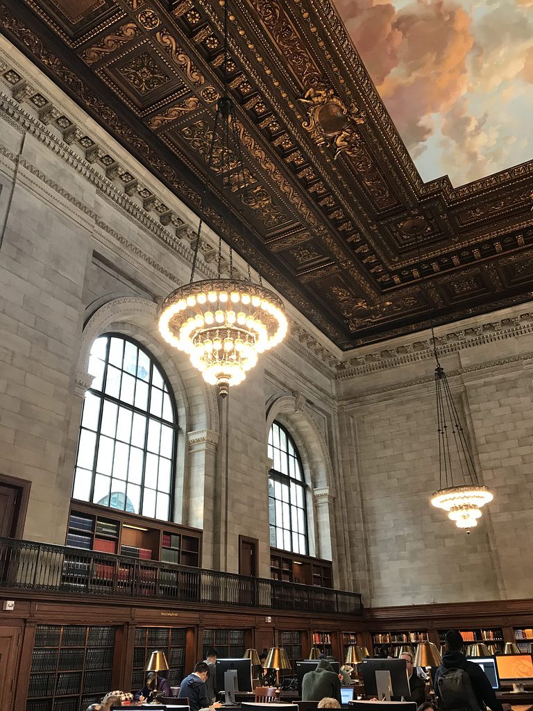 interior space in the New York Public Library showing several people studying, and French style paintings on the roof 