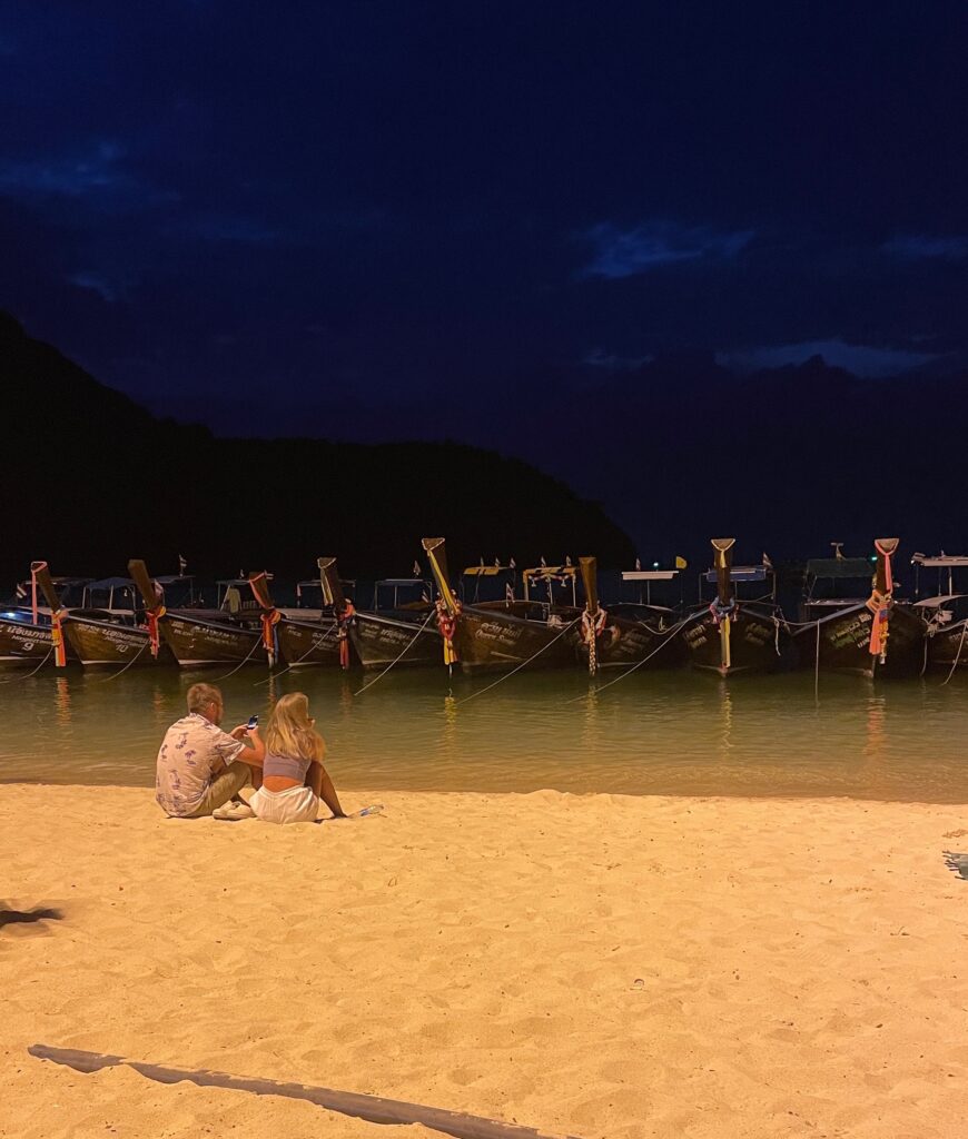 several longtail boats docked on the beach in Phi Phi, a couple sits under the lights on the beach