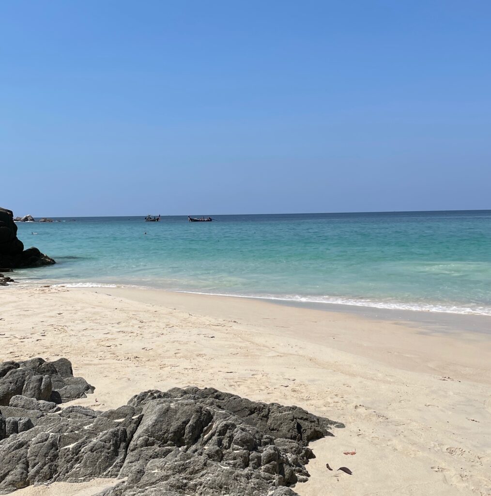 a few large rocks on a white sandy beach with stunning clear blue waters and long tail boats in the distance at Freedom Beach in Phuket