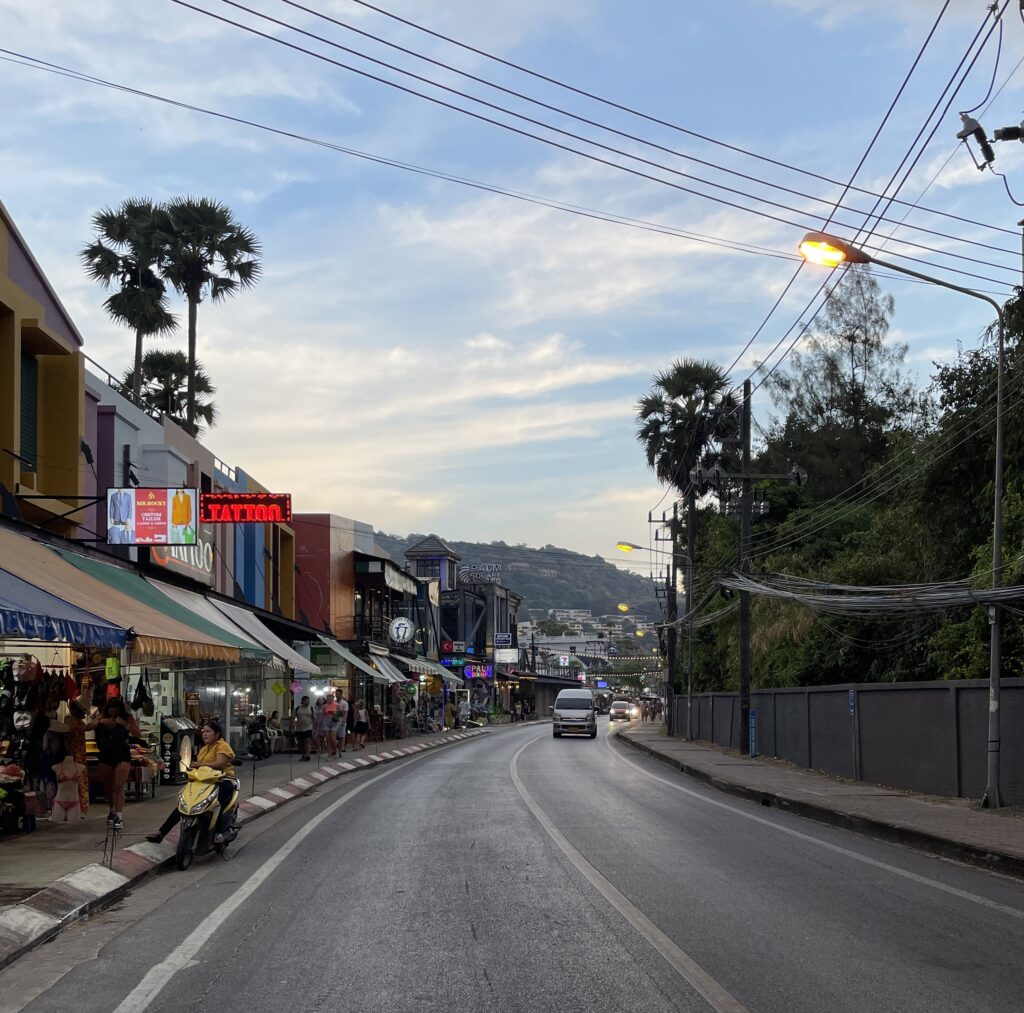 Several shops and tourist shopping, eating along the road as the sun is setting in with palm trees in the distance in Kata Beach, Phuket, Thailand