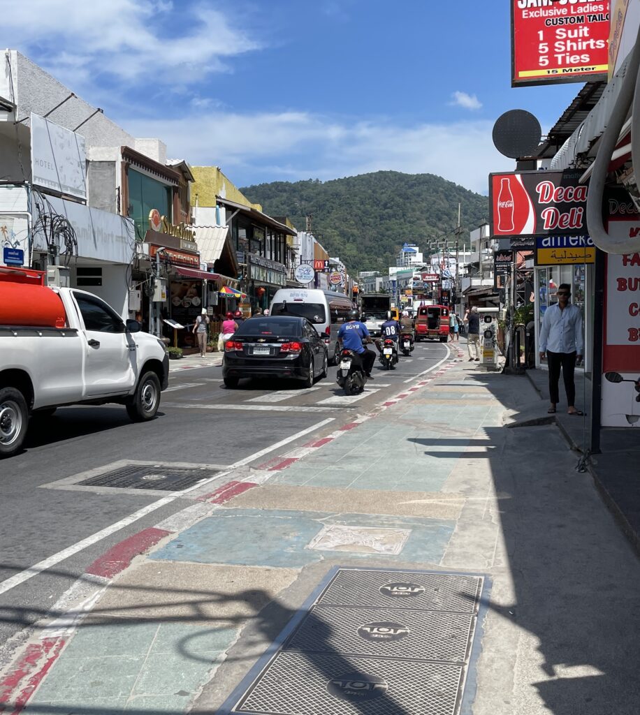 a road in Patong, Phuket with several cars, and clear skies and mountains in the distance