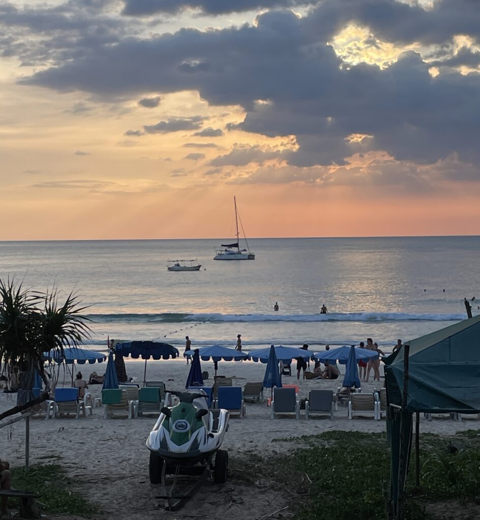 several people sitting on their beach lounge chairs watching the beautiful sunset and a sailboat passing by at Kata Beach in Phuket, Thailand