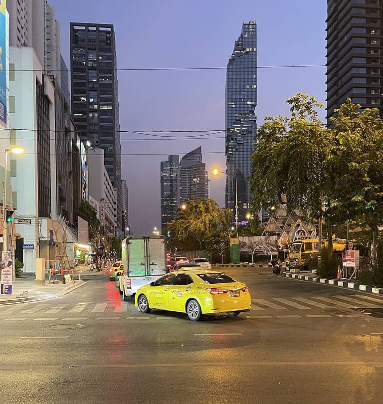 a yellow taxi passing by on a street at sunset with several high rise towers in the background in Bangkok, Thailand / is Bangkok Safe at night?