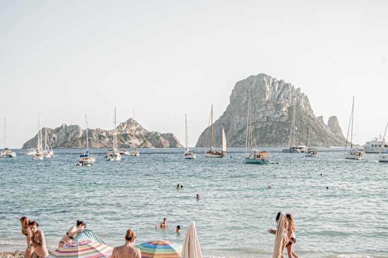 a few beachgoers walking along the picturesque shoreline amongst many boats in the water in Ibiza, Spain / Places like Ibiza to Visit