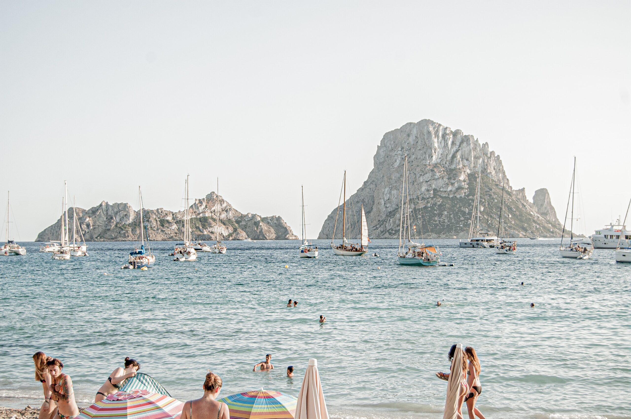 a few beachgoers walking along the picturesque shoreline amongst many boats in the water in Ibiza, Spain / Places like Ibiza to Visit