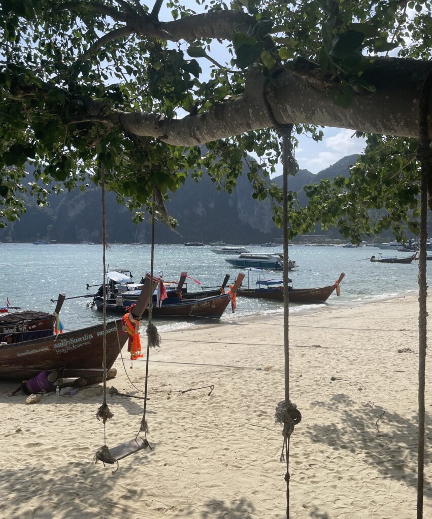 a wooden swing hanging on a tree with many Thai longtail boats on the shoreline on a beach in Koh Phi Phi in Krabi 