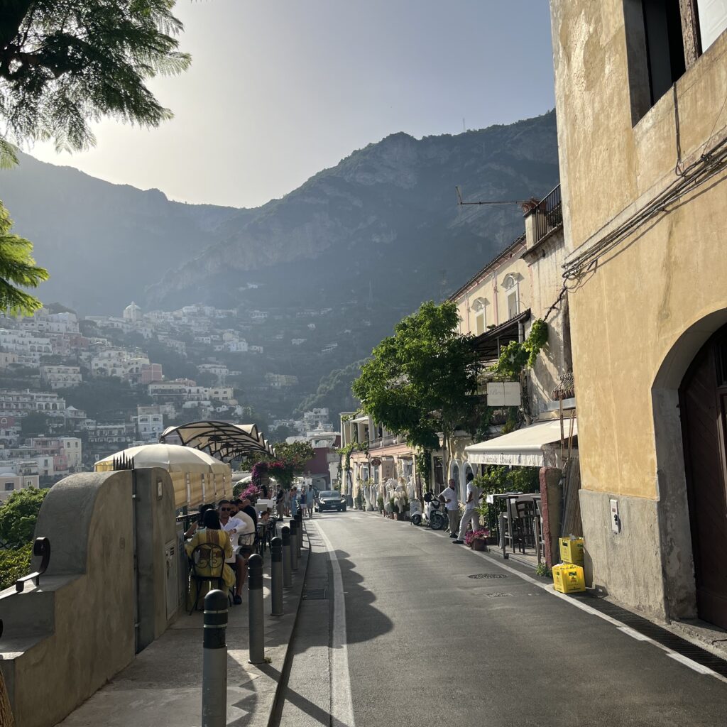 a picteresque street where locals and tourists are dining, hills are seen in the distance in  Positano, Italy 