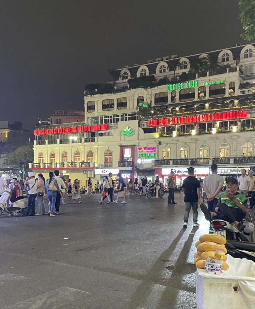 several locals and tourists walking in the night amongst many lit buildings on one of Hanoi's most famous streets