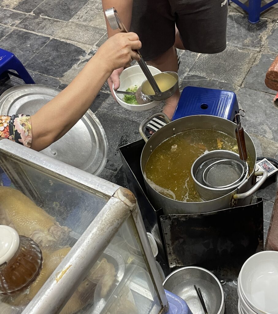 a woman pouring pho broth into a bowl for a man 