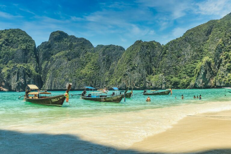 Traditional Thai longtail boats lined up on a sandy beach with dramatic limestone cliffs in the background.