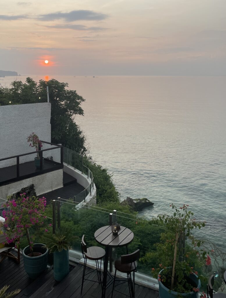 Sunset view from a cliffside beach club in Bali overlooking the ocean, with golden skies and waves crashing below.