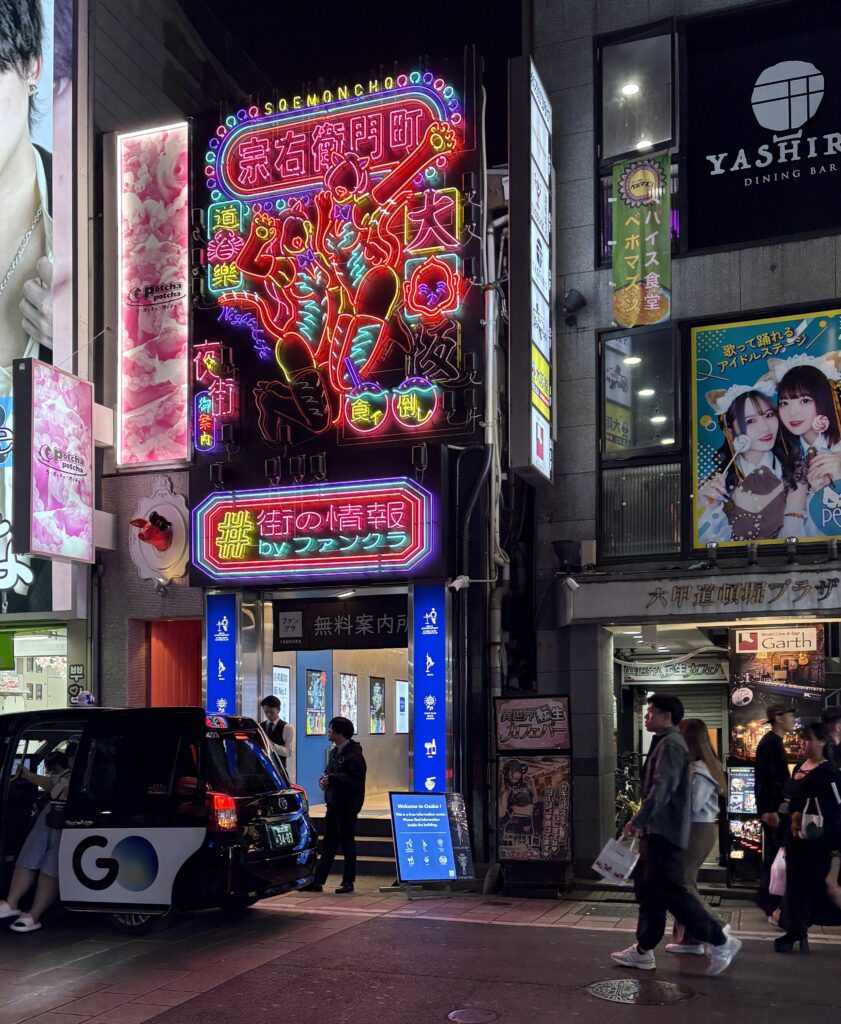People walking under colorful street signs and glowing advertisements in Osaka’s bustling nightlife district at night.