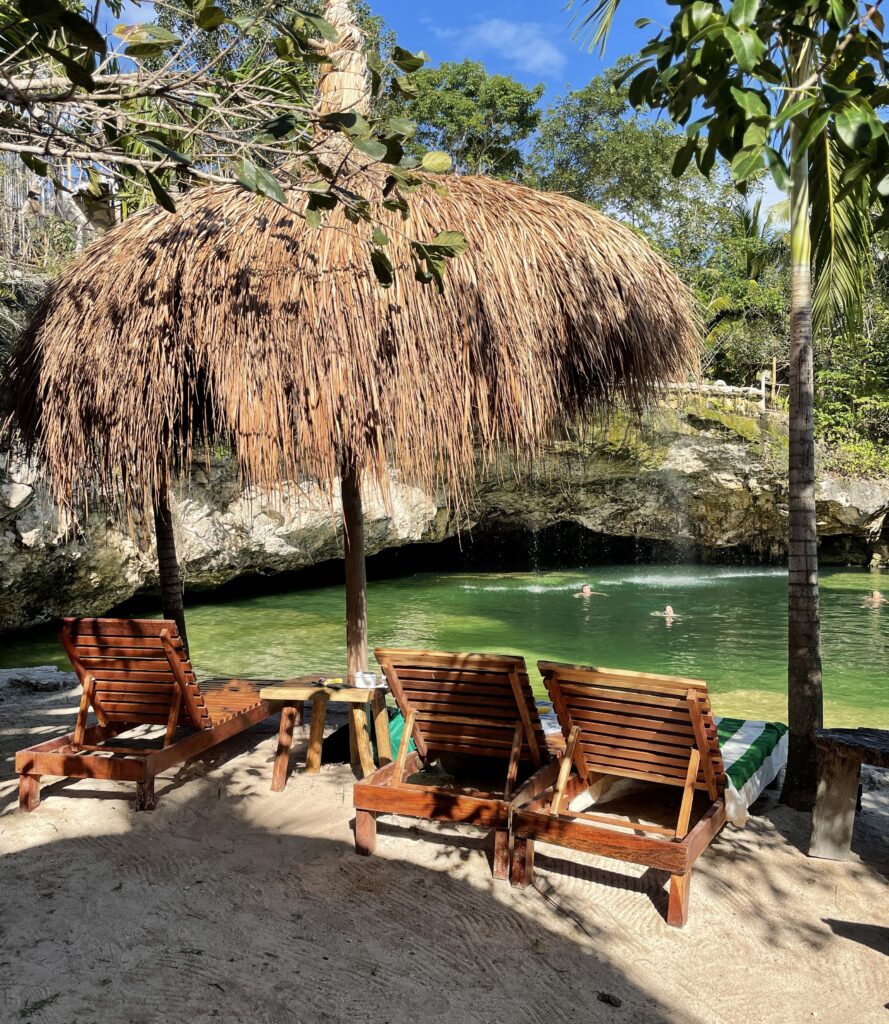 People swimming in a Tulum cenote with wooden beach chairs on the lawn under the sun