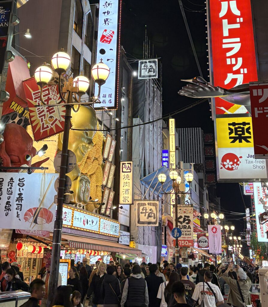 Crowds of people walking through a busy street in Dotonbori, Osaka, surrounded by shops and neon signs
