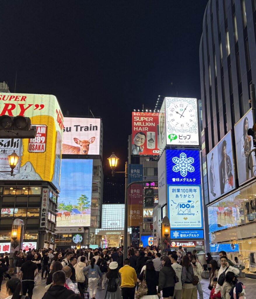 A bustling scene of people walking and shopping along Dotonbori’s neon-lit street in Osaka.
