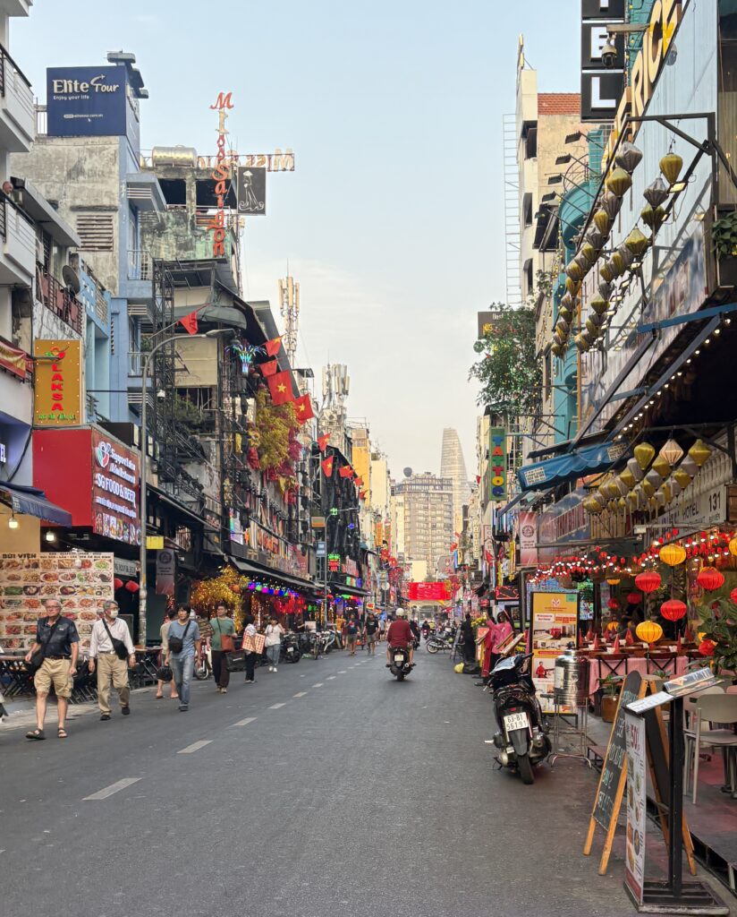 Early evening street in Ho Chi Minh City with sunlight still present, lined with restaurants, hotels, and shops, as scooters pass by.