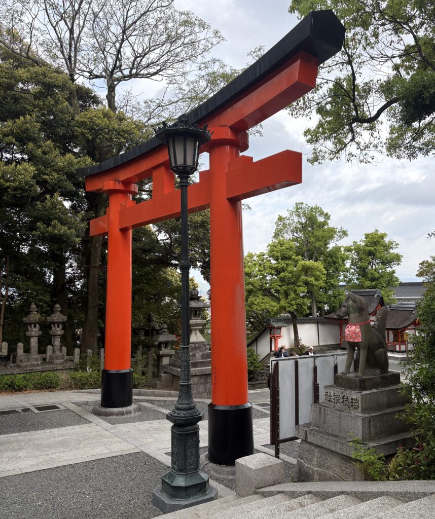 Red torii gate and dog statue at Fushimi Inari Taisha in Kyoto, captured from the stone stairs with early morning light and lush greenery.