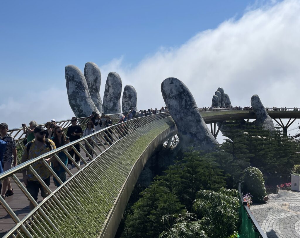 Tourists walking along the iconic Golden Bridge held by giant hands at Ba Na Hills, captured on a clear day in Da Nang.