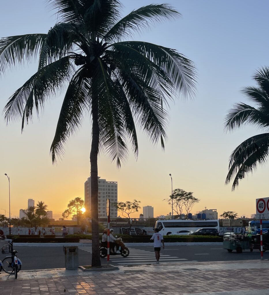A golden sunset over Da Nang as scooters pass by and a man crosses the road, with the city skyline visible in the distance, all viewed from the My Khe Beach boardwalk.