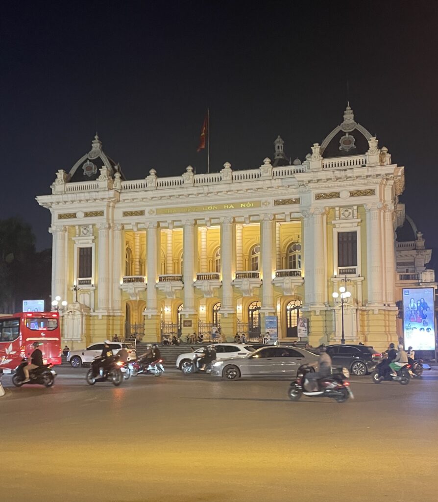 Hanoi Opera House illuminated at night with numerous scooters passing by in the French Quarter.