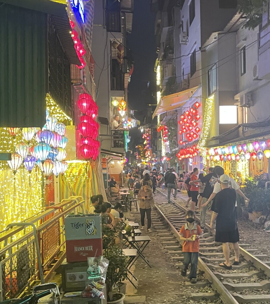 Locals and tourists walking on the train tracks in Hanoi at night with brightly lit stores and lanterns