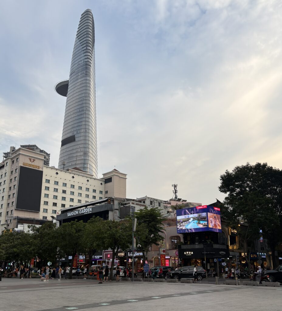 Main square in Ho Chi Minh City near sunset, with people walking along the pedestrian street and the tall skyscraper with a helipad visible in the background
