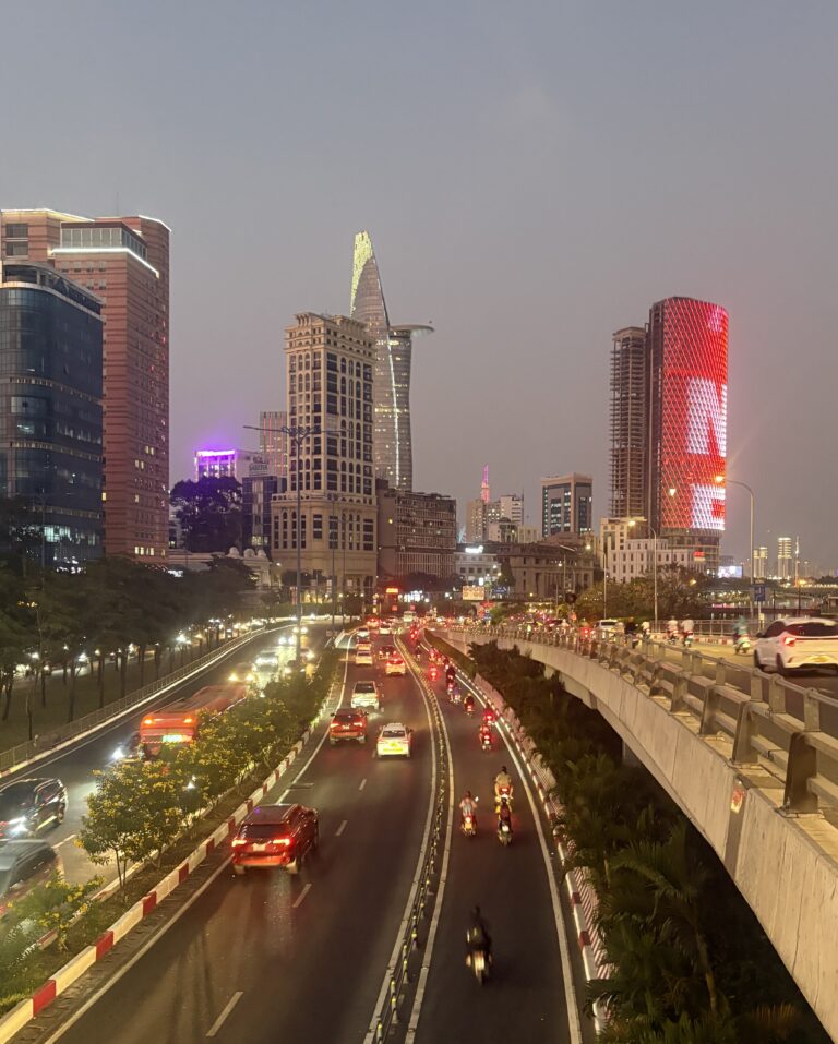 Evening view of a busy highway in Ho Chi Minh City at sunset, with cars passing and a distant building displaying the Vietnamese flag.