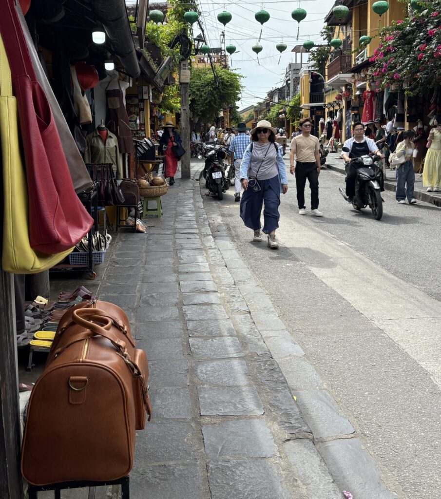 Evening in Hoi An Old Town with tourists strolling past a shop displaying a beige buffalo leather duffle bag on the left.