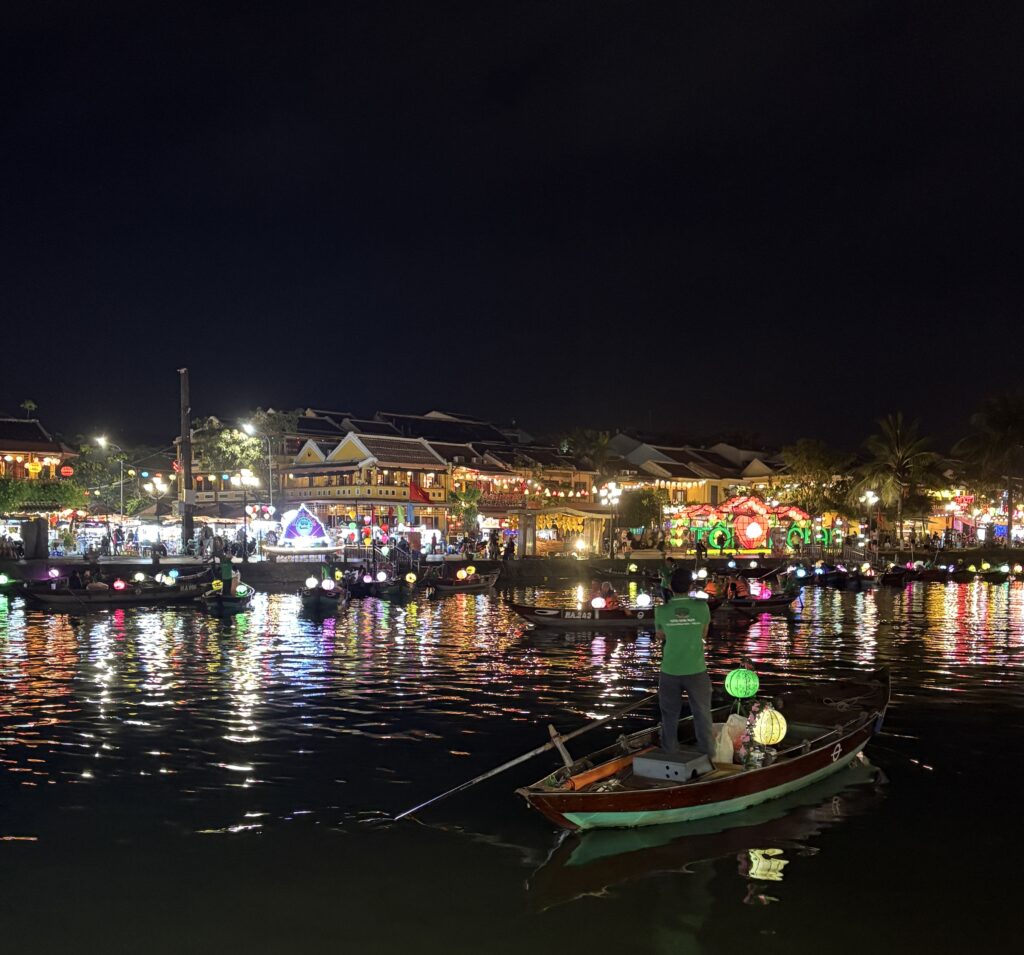 A man rows a small lantern-lit boat along the river as Hoi An Old Town glows with colorful lights and reflections at night.