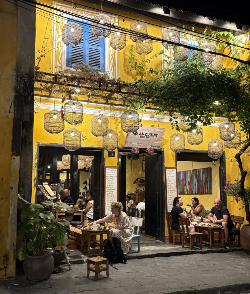 Tourists dining at a restaurant inside a charming yellow colonial building in Hoi An Old Town at night.
