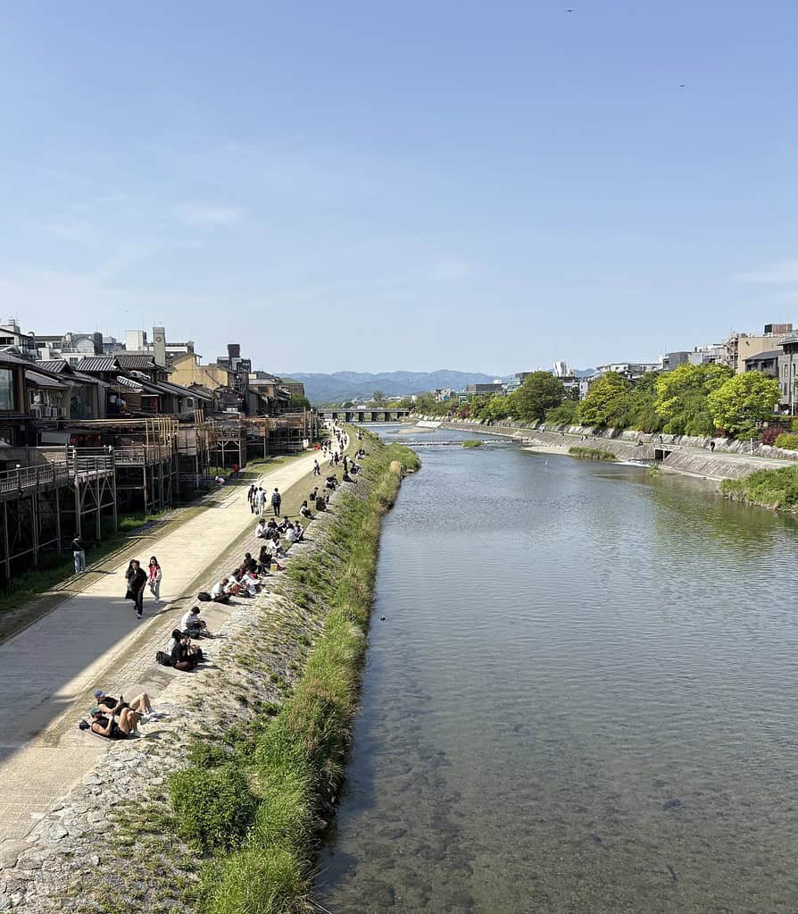 Locals and tourists relaxing by the Kamogawa River in Kyoto with distant mountains on a sunny day
