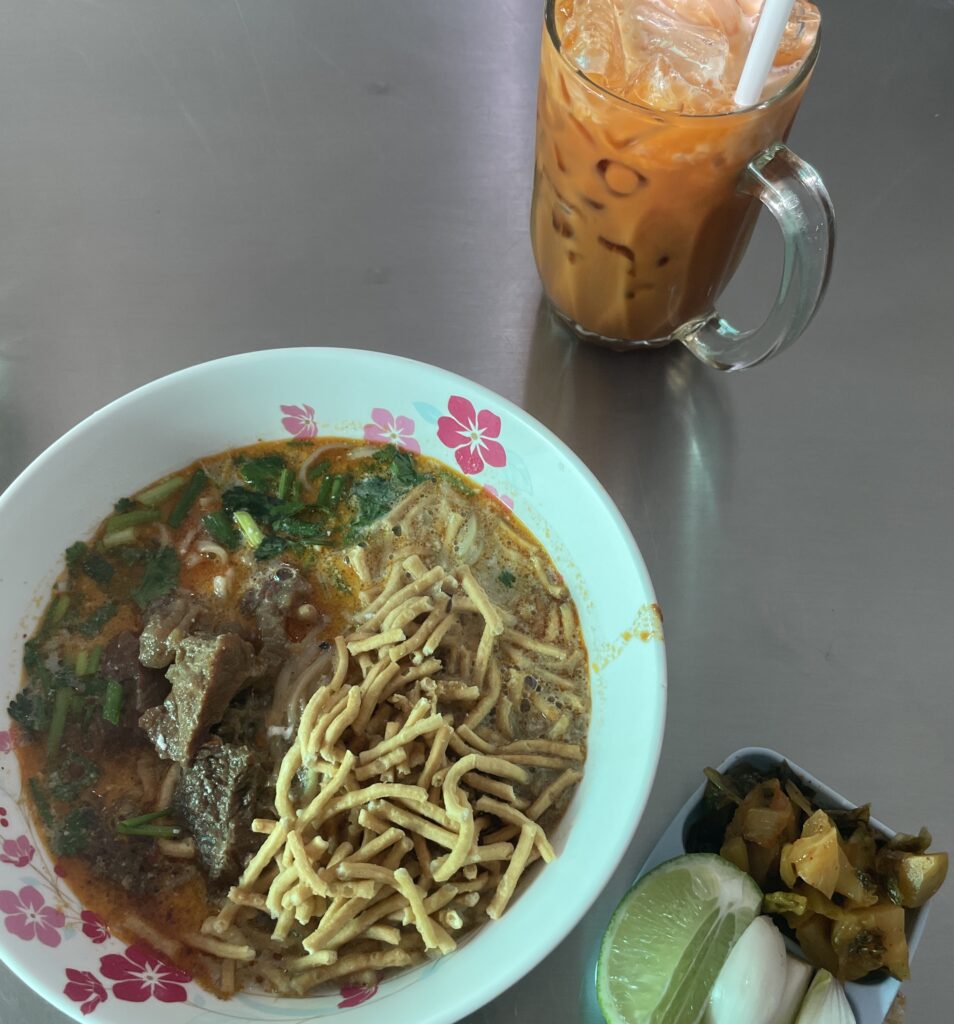 Beef Khao Soi served at Khao Soi Islam in Chiang Mai, with a side of traditional noodles and a glass of Thai milk tea in the background.