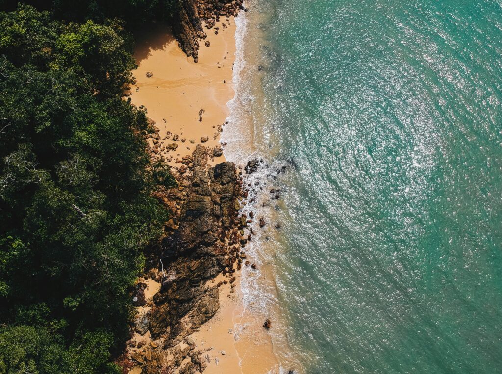 Aerial view of a remote beach with rocky shoreline, dense greenery, and turquoise water in Koh Phayam, Thailand