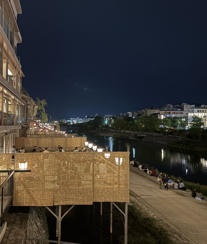 Locals and tourists sitting at riverside cafes in Kyoto during the evening, enjoying a laid-back atmosphere along the water.