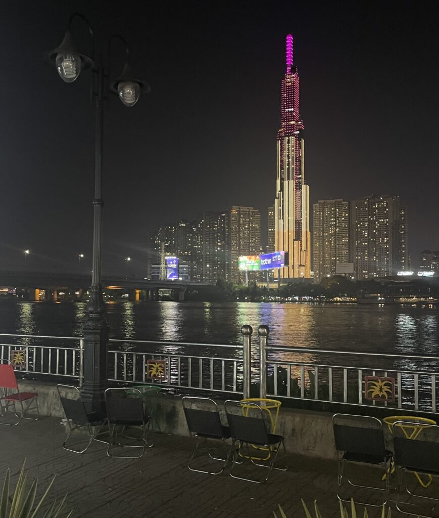 Brightly lit Landmark 81 building in Ho Chi Minh City with a river view and seating area by the water at night.
