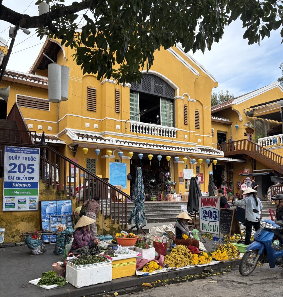 Vietnamese street vendors wearing traditional hats selling colorful flowers on the ground with yellow-painted UNESCO buildings in the background in Hoi An Old Town.