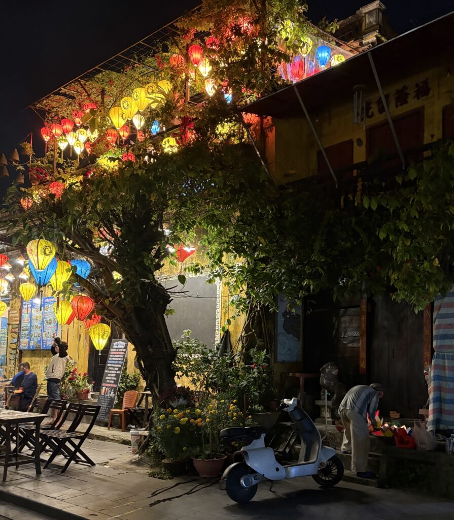 Local woman kneeling to light small lantern boats in Hoi An Old Town at night, with a lantern-filled restaurant glowing in the background