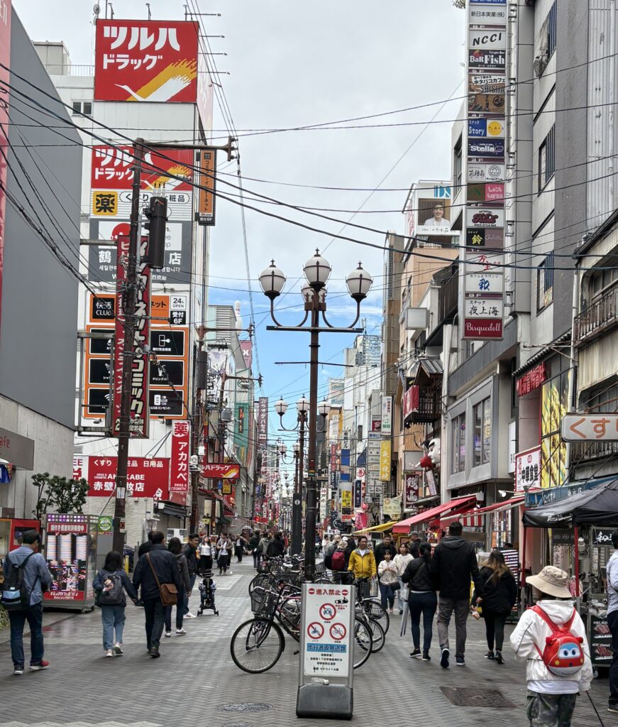 Locals and tourists walking along one of the main streets in Dotonbori, Osaka, during the day, surrounded by shops and vibrant signage.