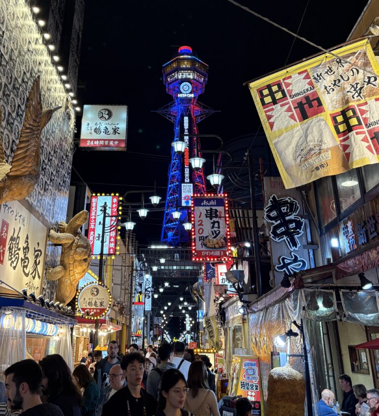 Locals and tourists strolling along Shinsekai’s main market street with Osaka’s illuminated Hitachi Tower glowing in the background