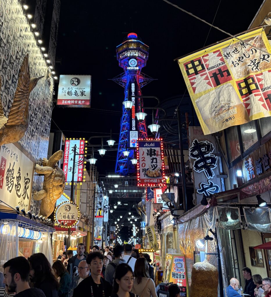Locals and tourists strolling along Shinsekai’s main market street with Osaka’s illuminated Hitachi Tower glowing in the background