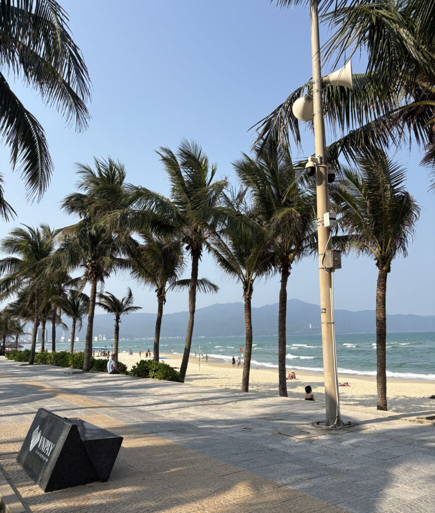 Early evening view of the My Khe Beach boardwalk in Da Nang, featuring palm trees, sandy beach, and mountains in the distance.