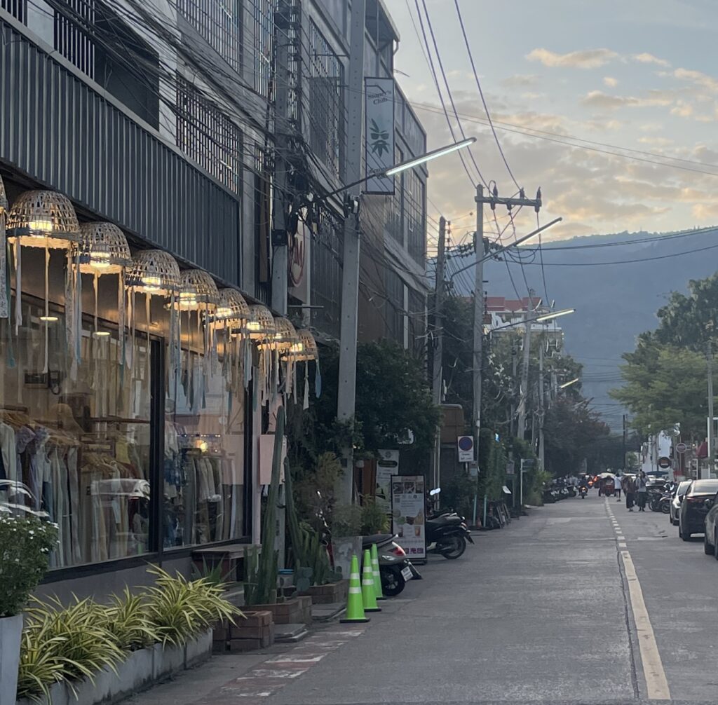 Street view in Nimman, Chiang Mai, with mountains in the distance at dusk.
