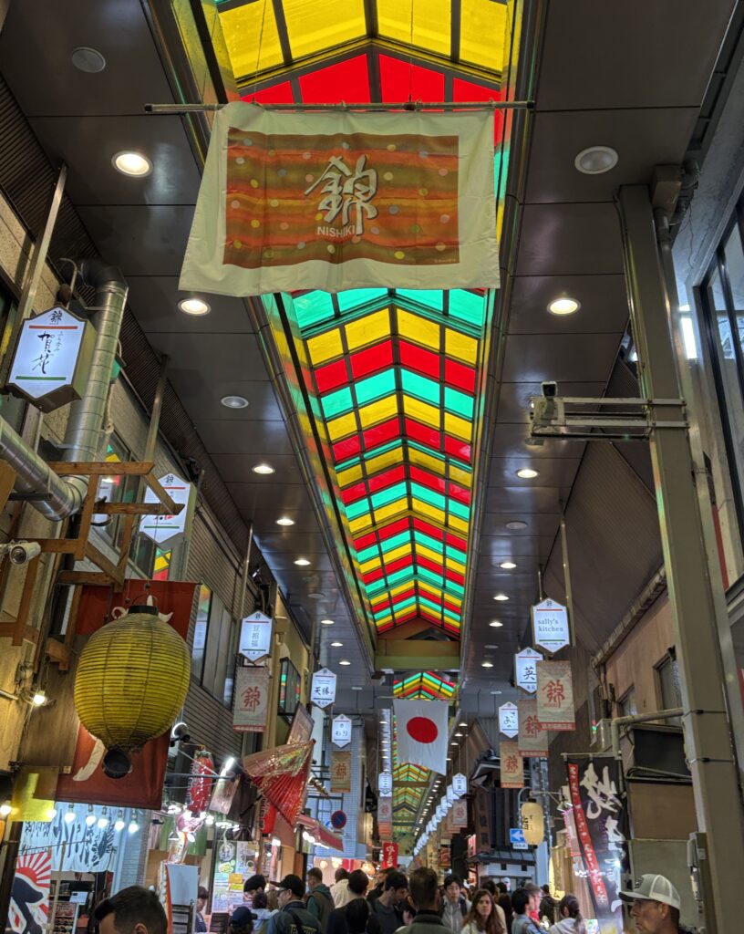 Tourists walking through Kyoto’s Nishiki Market under the colorful covered roof, browsing local stalls and shops