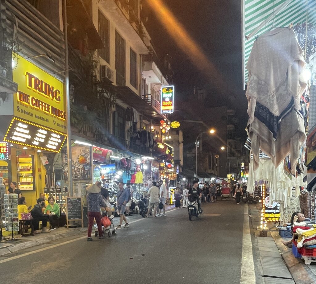 A lively street in Hanoi’s Old Quarter at night, lined with shops selling clothing, souvenirs, and local goods.