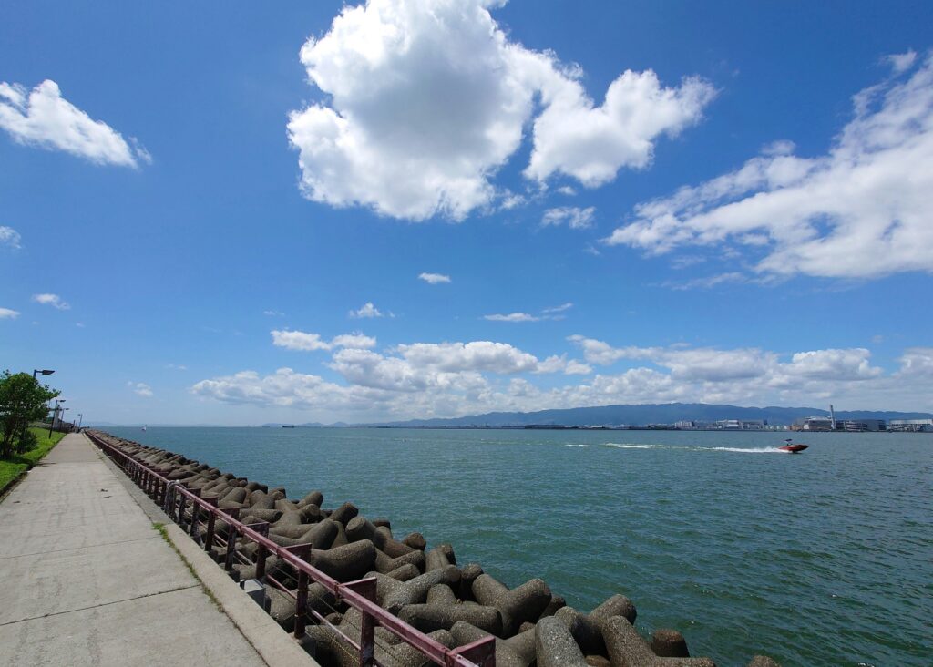 Scenic view of Osaka Bay Marina with a boat passing by and people walking along the sunny waterfront promenade.
