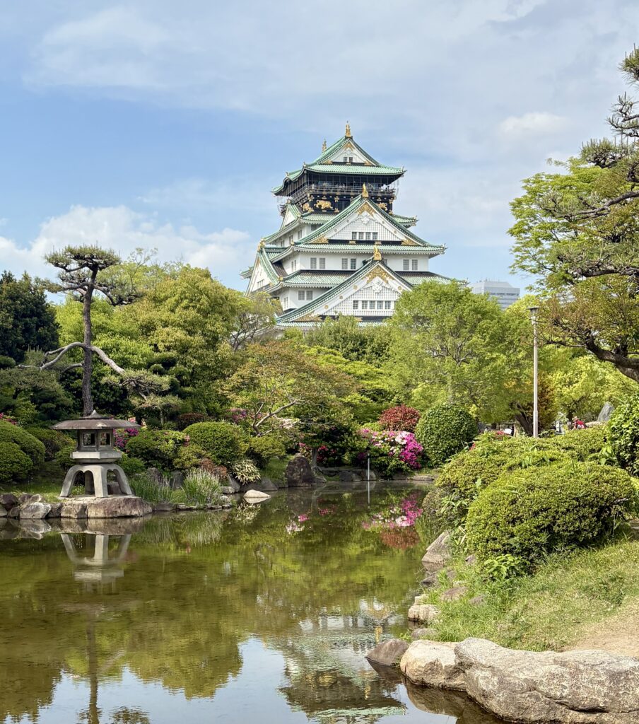 Osaka Castle rising behind a tranquil garden and pond, captured on a clear sunny day with lush greenery and reflection in the water