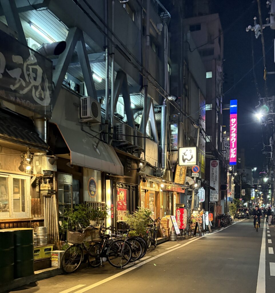 A cozy side street in Osaka lined with lit-up restaurants, with a local man biking through the evening ambiance.