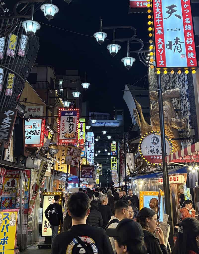 Tourists walking through a busy street at night in Dotonbori, Osaka, surrounded by neon signs and nightlife energy.